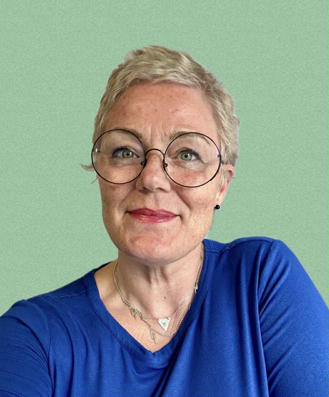 Porträtt av Pernilla Pernsjö med kort, blont hår och glasögon, iklädd en blå tröja, poserande framför en grön bakgrund.